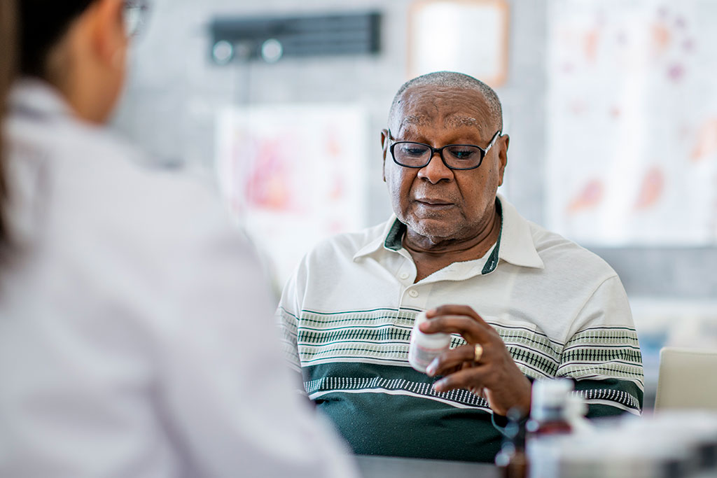 An elderly man looks at a pill bottle in his hand while sitting across the table from a doctor in the doctor's office. He appears to be making a decision about it.